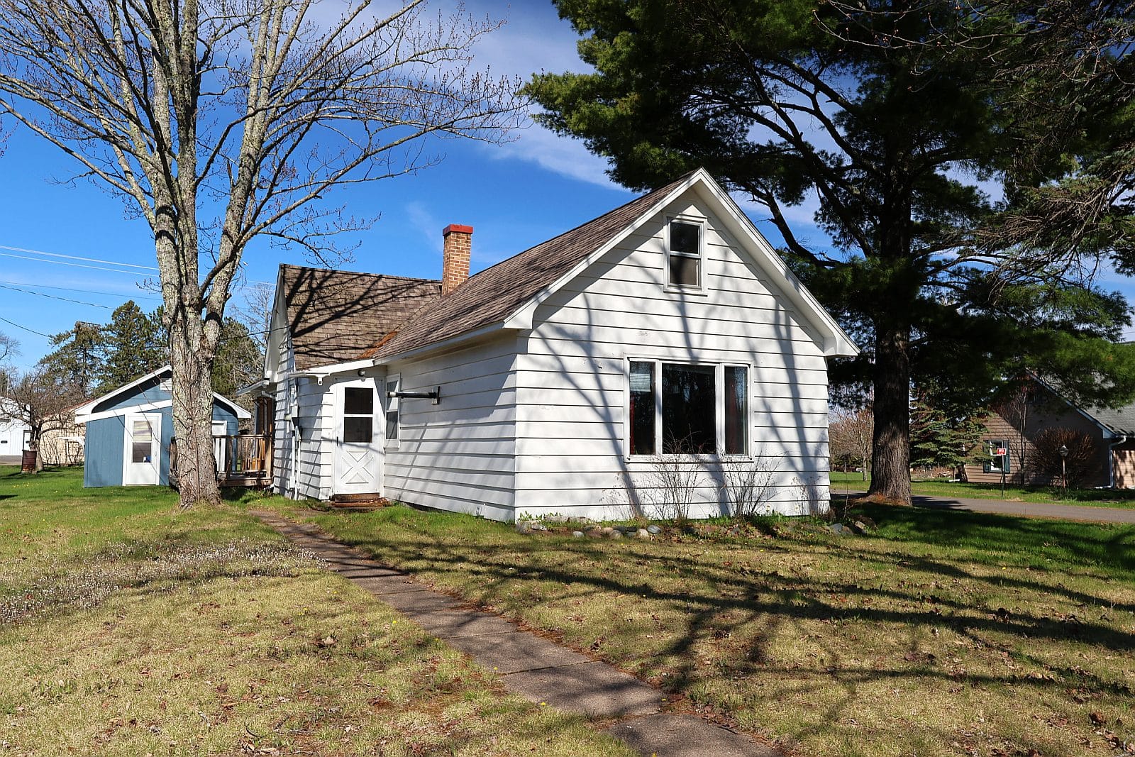 83185 Washington Ave, Port Wing, WI - 1.5-story white-sided single family home with steep front gable, brick chimney, upper gable window, mature trees, and a concrete walkway to the side entry on a corner lot