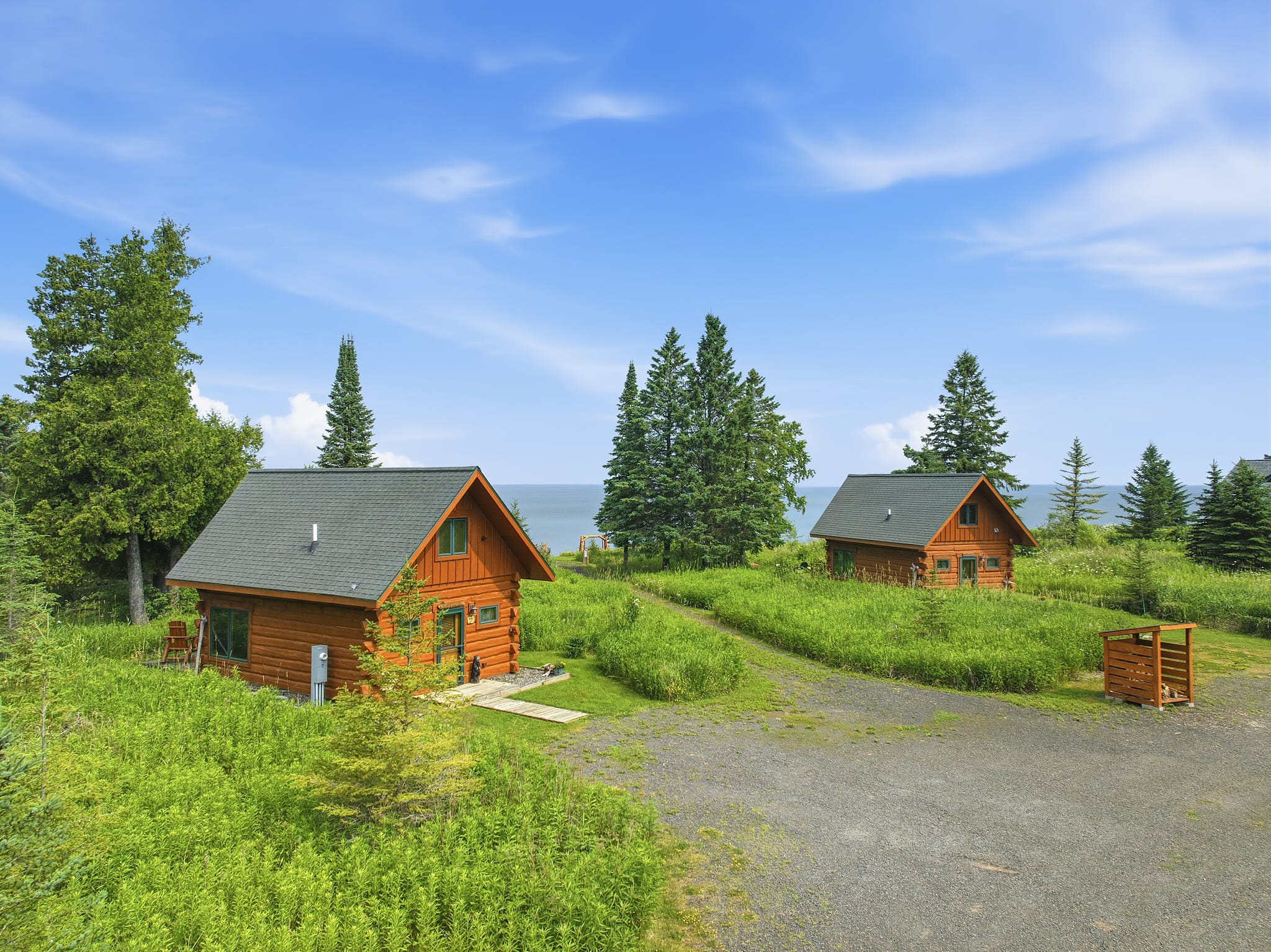 Aerial view of two log cabins with green metal roofs set in a grassy meadow above Lake Superior's south shore in Port Wing, WI, with a gravel driveway connecting the two homes and mature evergreen trees framing the view. FOR SALE.