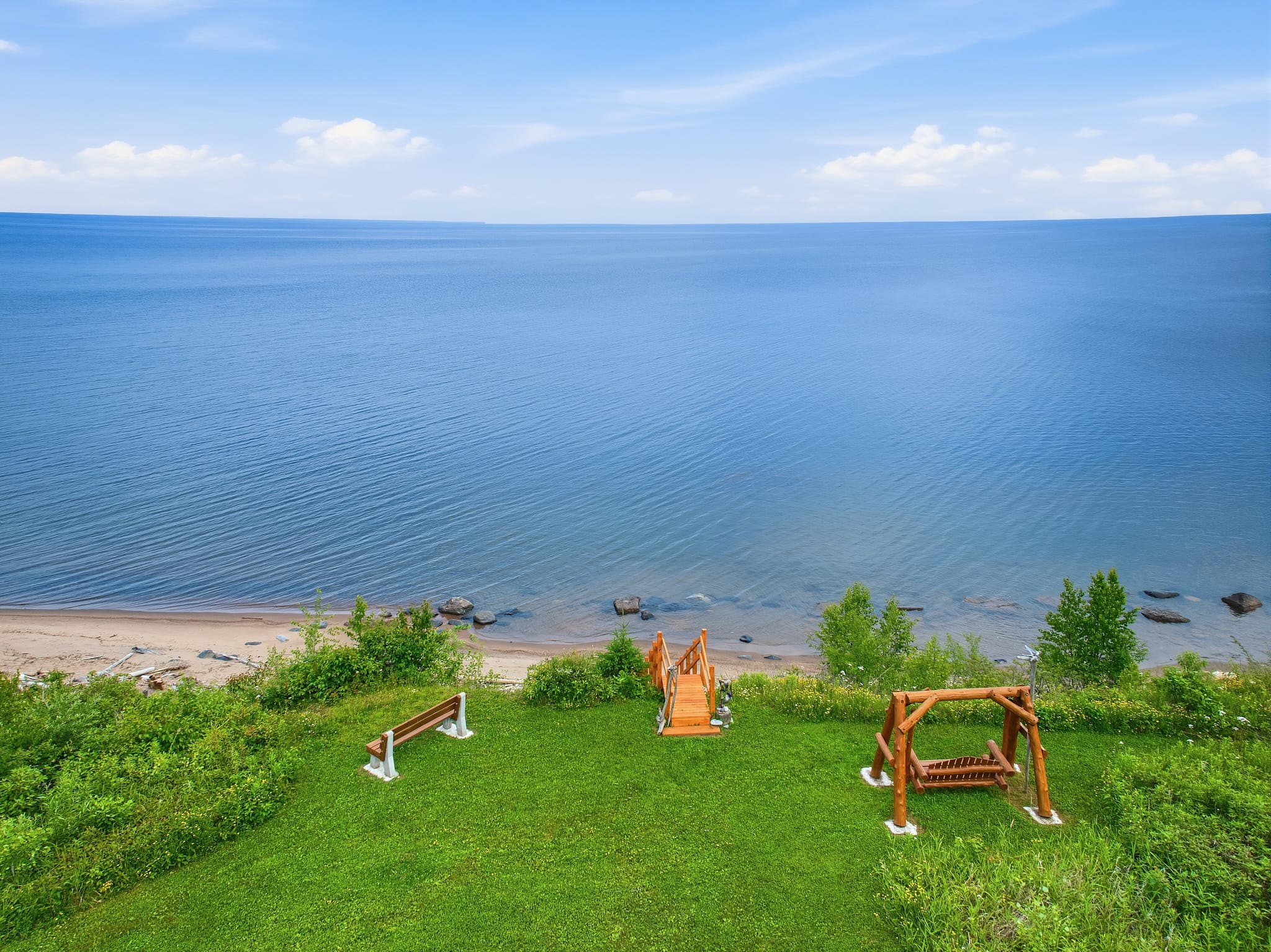 Aerial view looking down a grassy bluff toward Lake Superior, showing a bench, log swing, and fire pit with Adirondack chairs arranged along a mowed path lined with evergreen trees.