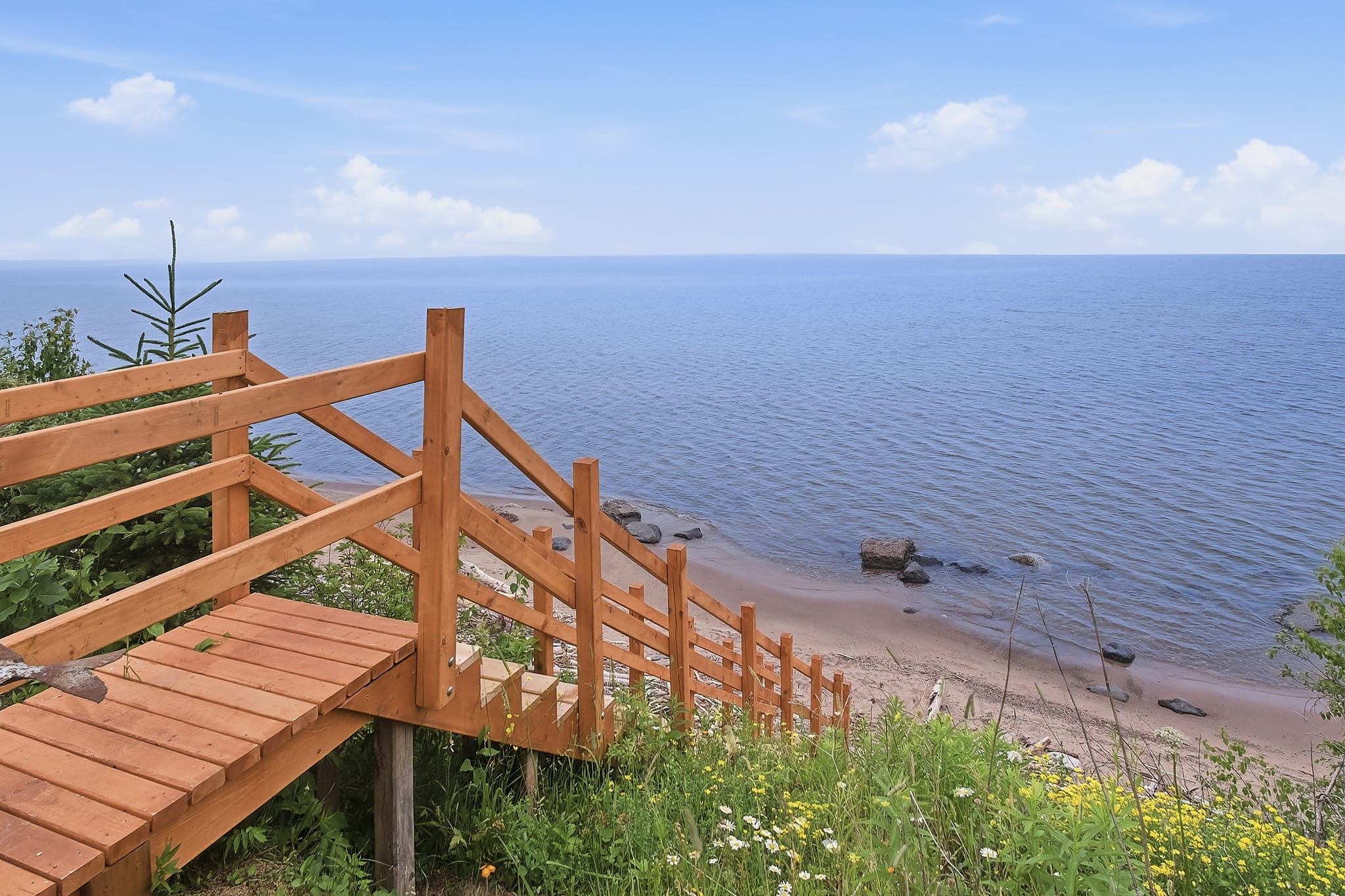 Wooden staircase with railings descending a grassy bluff to a sandy Lake Superior beach with scattered rocks, under a blue sky with scattered clouds.