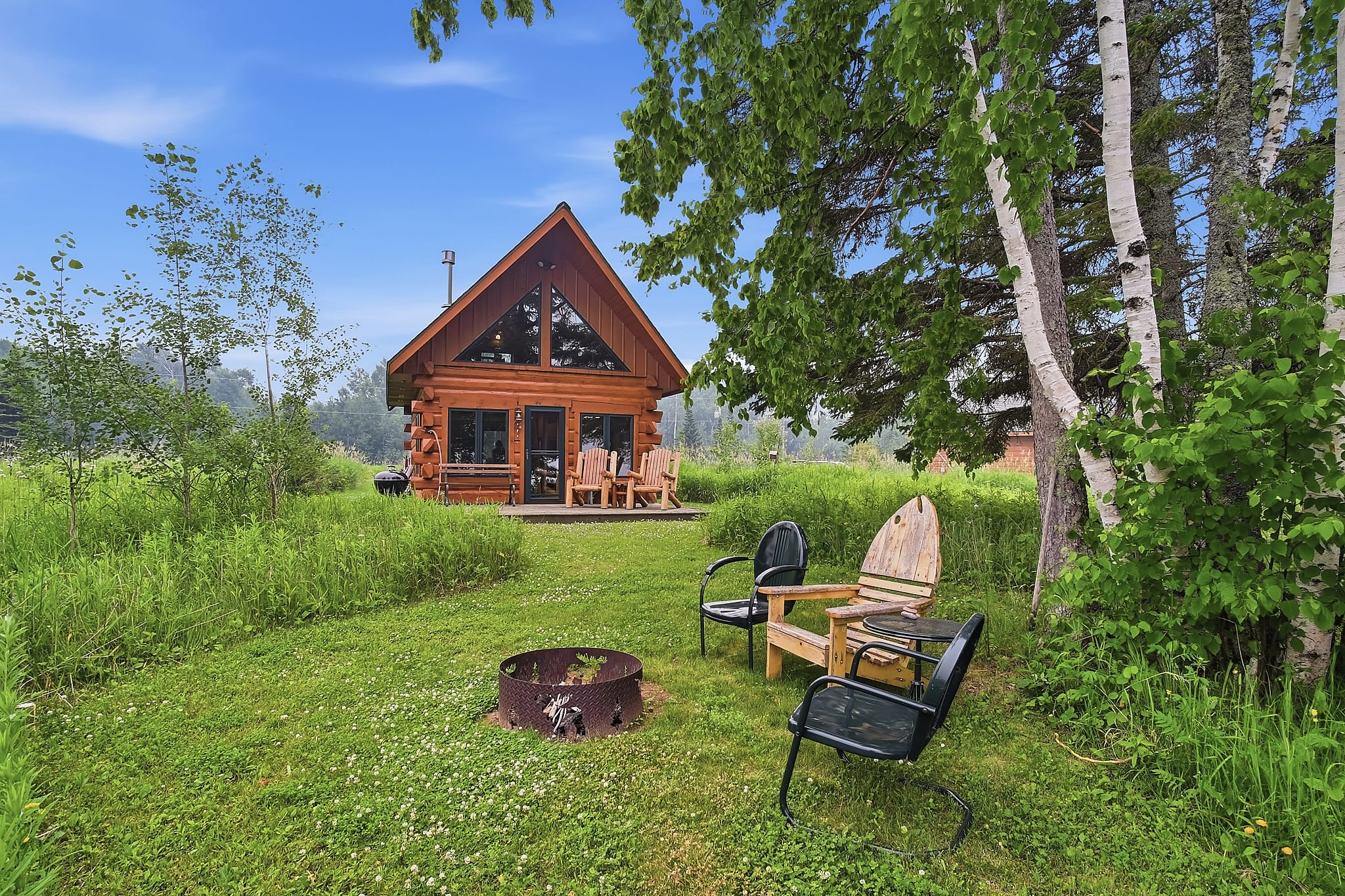 Two Adirondack chairs around a metal fire pit on a gravel patio overlooking a mowed lawn, a bench, a log swing, and Lake Superior in the distance.
