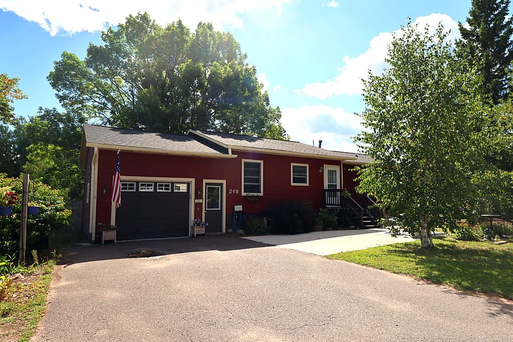 Red ranch home for sale at 218 E Bayfield St in Washburn, Wisconsin, photographed from the end of the asphalt driveway on a sunny day, with an attached single-car garage with a dark overhead door and window panels, cream-trimmed windows, house number 218 beside the front door, an American flag mounted by the garage, a covered side entry with wood steps and railing, a concrete walk to the entry, flower planters, and a white birch and mature shade trees framing the lawn