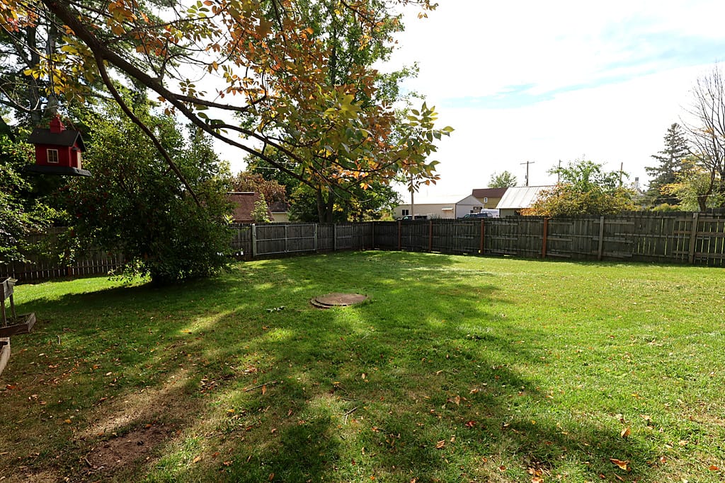Fenced backyard at 218 E Bayfield St in Washburn, Wisconsin, with a level mowed lawn, a covered brick firepit set in the grass, a wood privacy fence with a metal double gate opening to the alley, a red birdhouse hanging from a shade tree, and a crabapple tree heavy with red fruit at the left edge