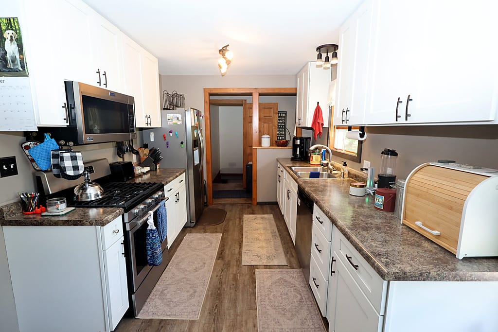 Galley kitchen at 218 E Bayfield St in Washburn, Wisconsin, with white shaker cabinets, dark speckled laminate countertops, a stainless gas range with over-range microwave, stainless French door refrigerator, stainless dishwasher, a double sink under a window, and wood-look vinyl plank flooring running to a doorway at the far end