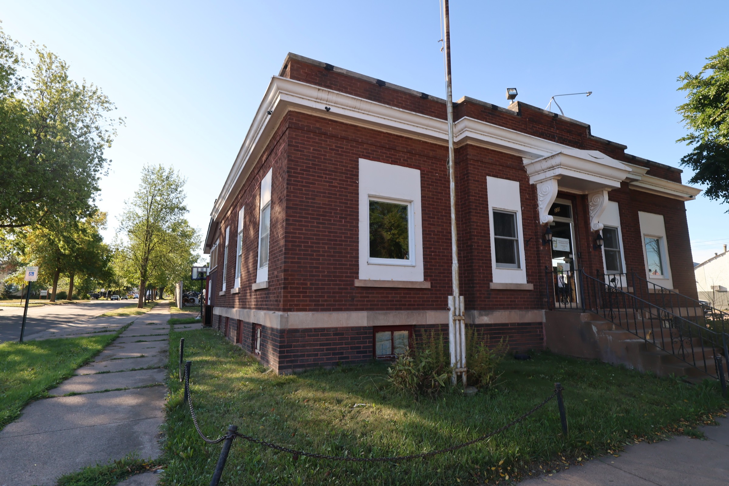 Brick commercial building for sale at 220 Main St E in downtown Ashland, Wisconsin, seen from the corner of Main Street and Third Avenue East, with red brick walls, a dark brick and stone base course, white-trimmed windows, a columned front entry with concrete steps and iron railings, and public sidewalk running along both street frontages